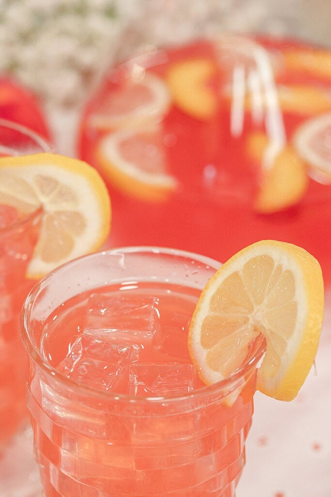 Close-up of a glass of homemade peach lemonade served over ice with a fresh lemon slice garnish, with a pitcher of peach lemonade in the background.