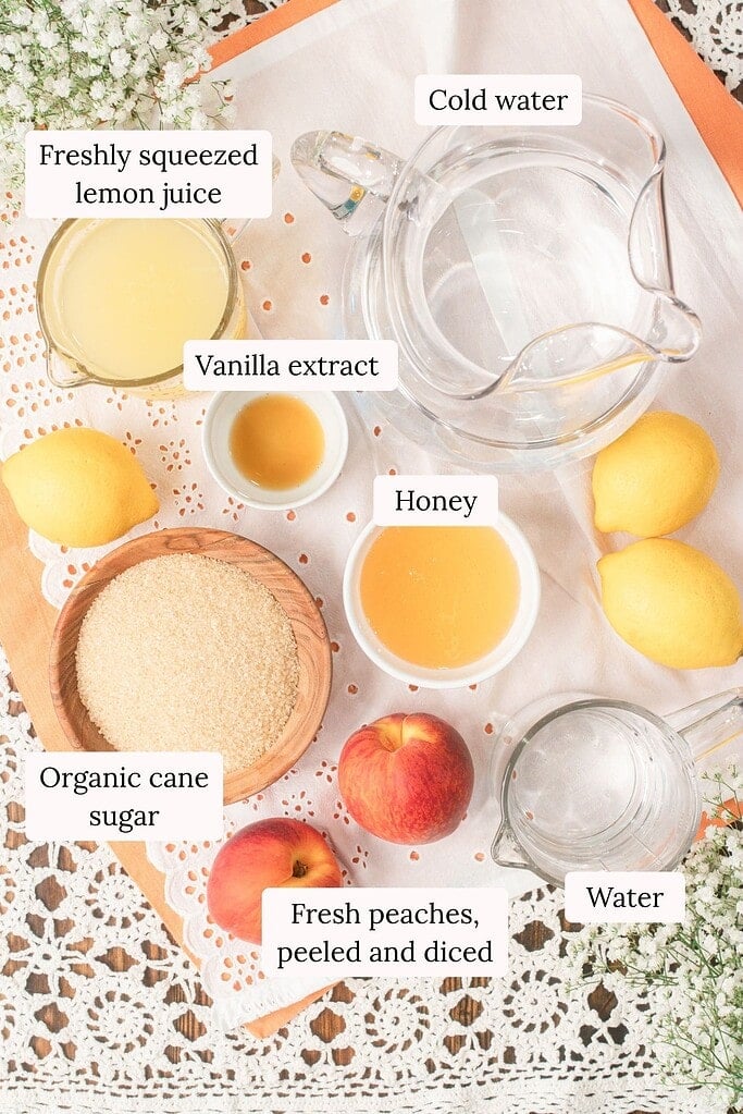 Overhead ingredient photo for homemade peach lemonade showing fresh peaches, organic cane sugar, honey, vanilla extract, fresh lemon juice, cold water, and water in glass pitchers on a linen cloth.
