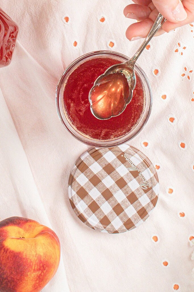 A spoon lifting homemade peach simple syrup from a glass mason jar, showing the smooth, glossy syrup made with fresh peaches and organic cane sugar.
