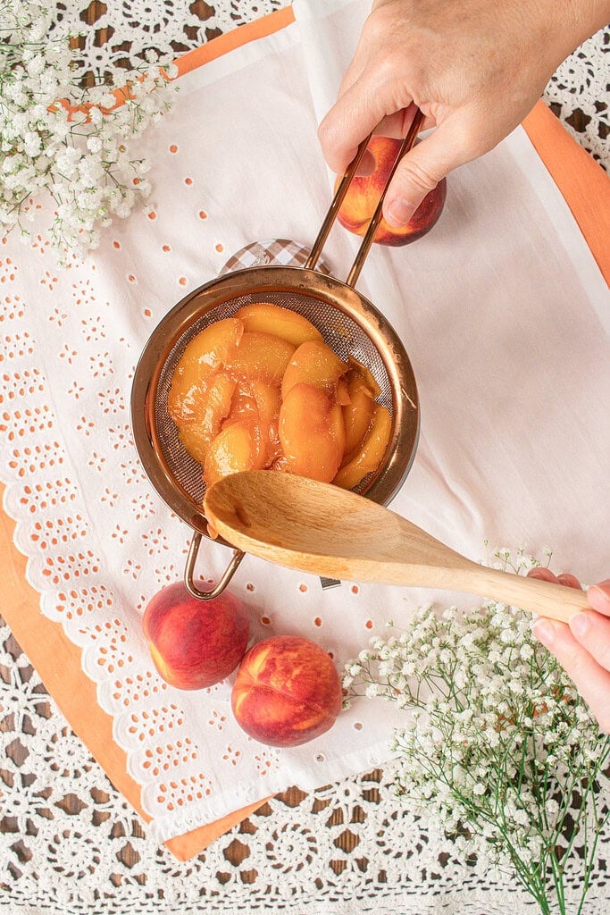 Straining cooked peaches through a fine-mesh sieve after making homemade peach simple syrup, while pressing the fruit with a wooden spoon to extract every drop of syrup.