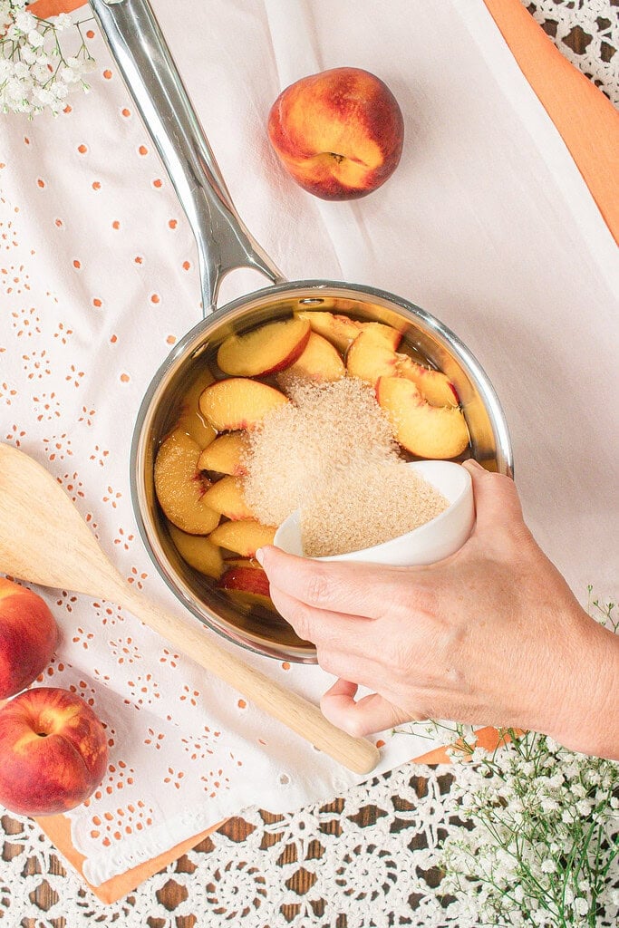 Adding organic cane sugar to sliced fresh peaches in a saucepan to make homemade peach simple syrup from scratch.