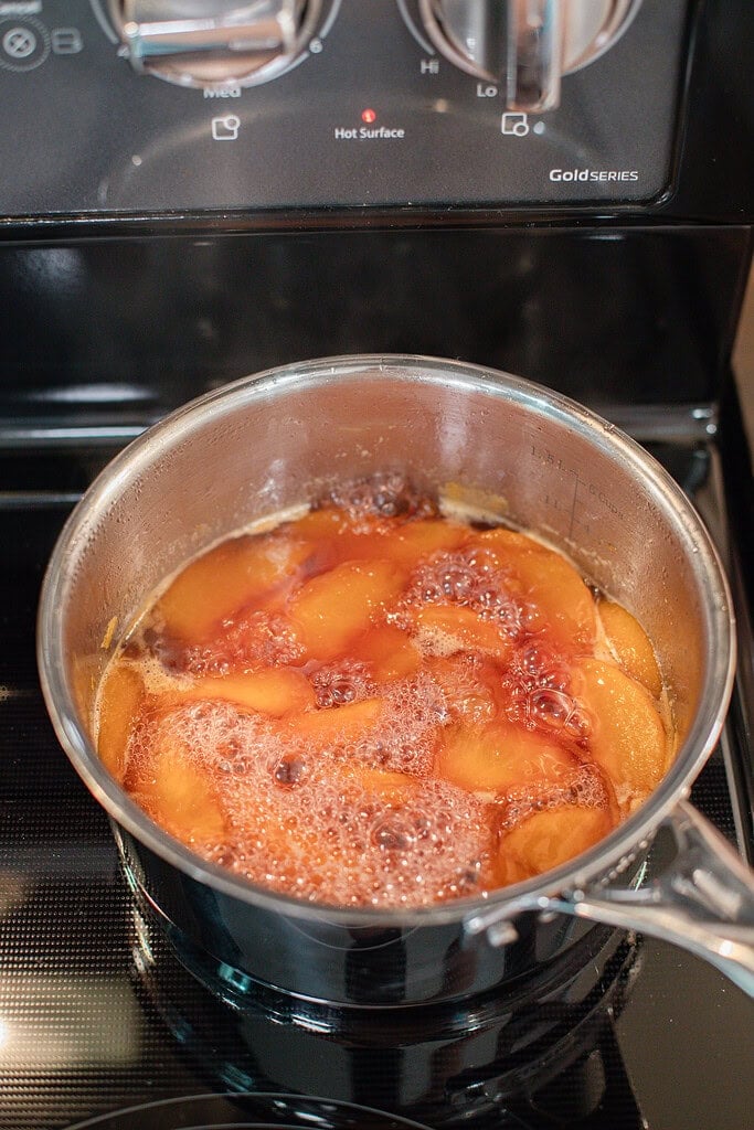 Fresh peach slices simmering in a saucepan with sugar and water while cooking homemade peach simple syrup on the stovetop.