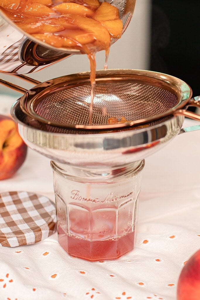 Pouring homemade peach simple syrup through a fine-mesh strainer into a glass mason jar, separating the smooth syrup from the cooked peaches.