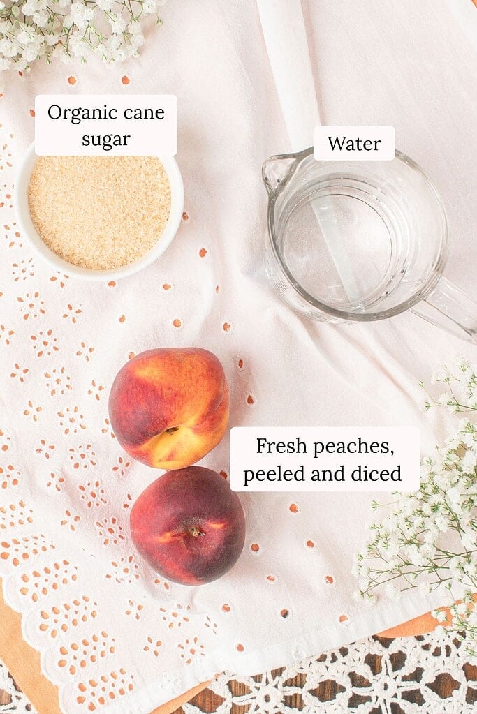 Ingredient photo for homemade peach simple syrup showing fresh peaches, organic cane sugar, and water arranged on a soft linen cloth with white flowers for an easy summer syrup recipe.