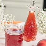 Homemade peach simple syrup stored in a glass mason jar beside a decorative glass bottle, surrounded by fresh peaches for serving and storage.