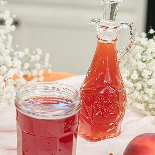 Homemade peach simple syrup stored in a glass mason jar beside a decorative glass bottle, surrounded by fresh peaches for serving and storage.