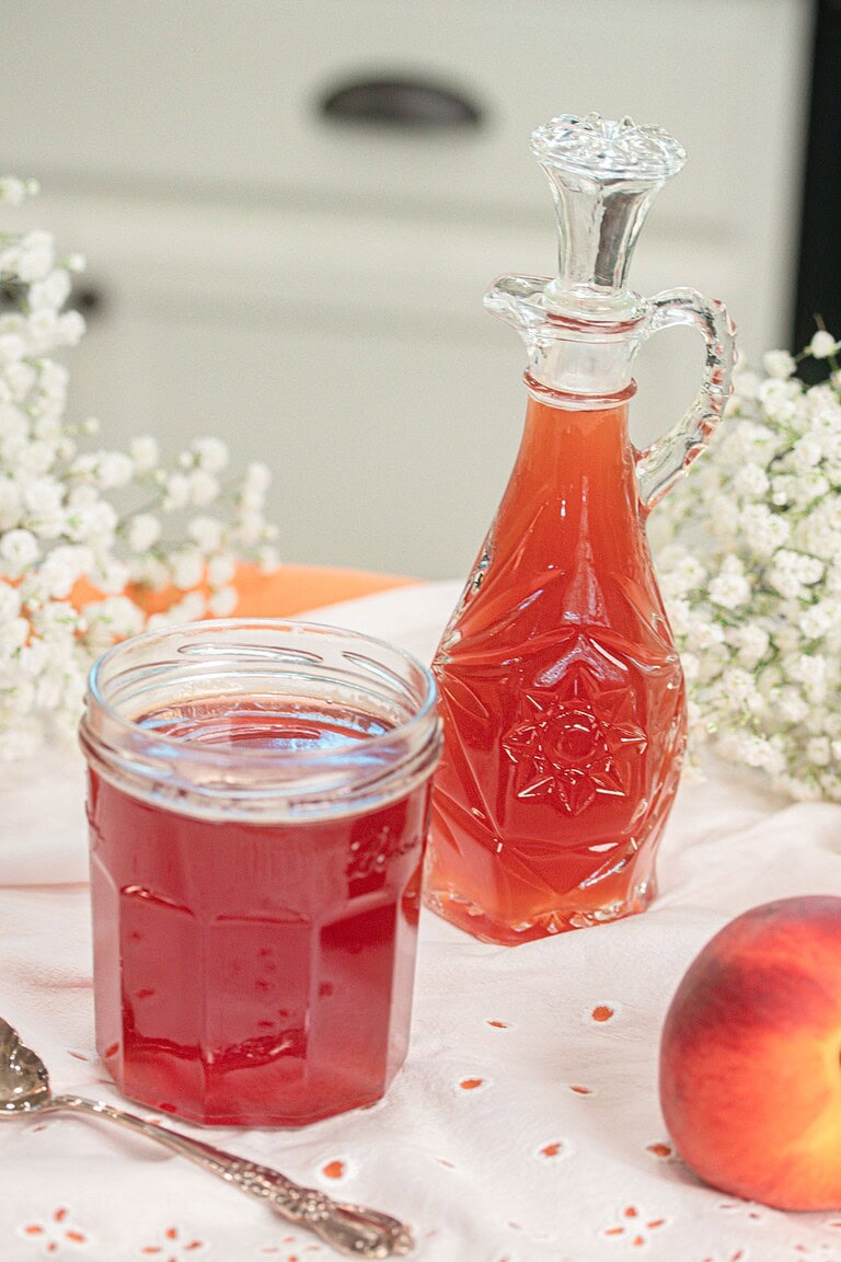 Homemade peach simple syrup stored in a glass mason jar beside a decorative glass bottle, surrounded by fresh peaches for serving and storage.