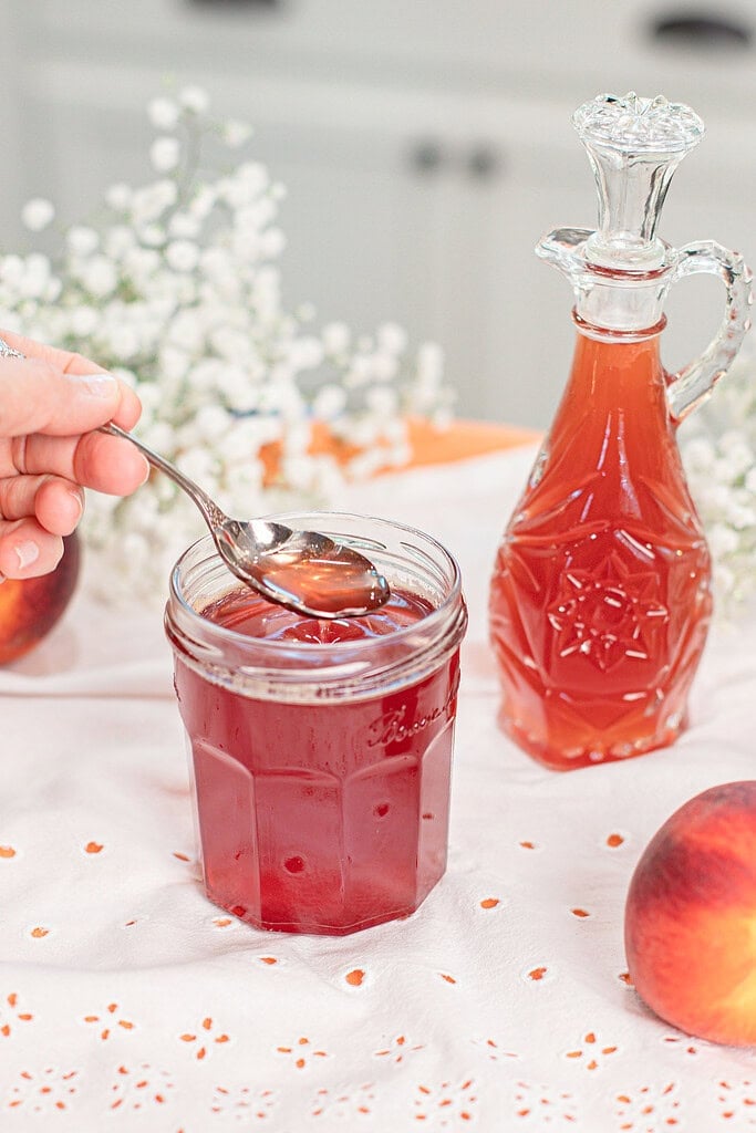 A spoon drizzling homemade peach simple syrup from a glass mason jar, showing the smooth consistency of the fresh peach syrup beside a decorative glass bottle.