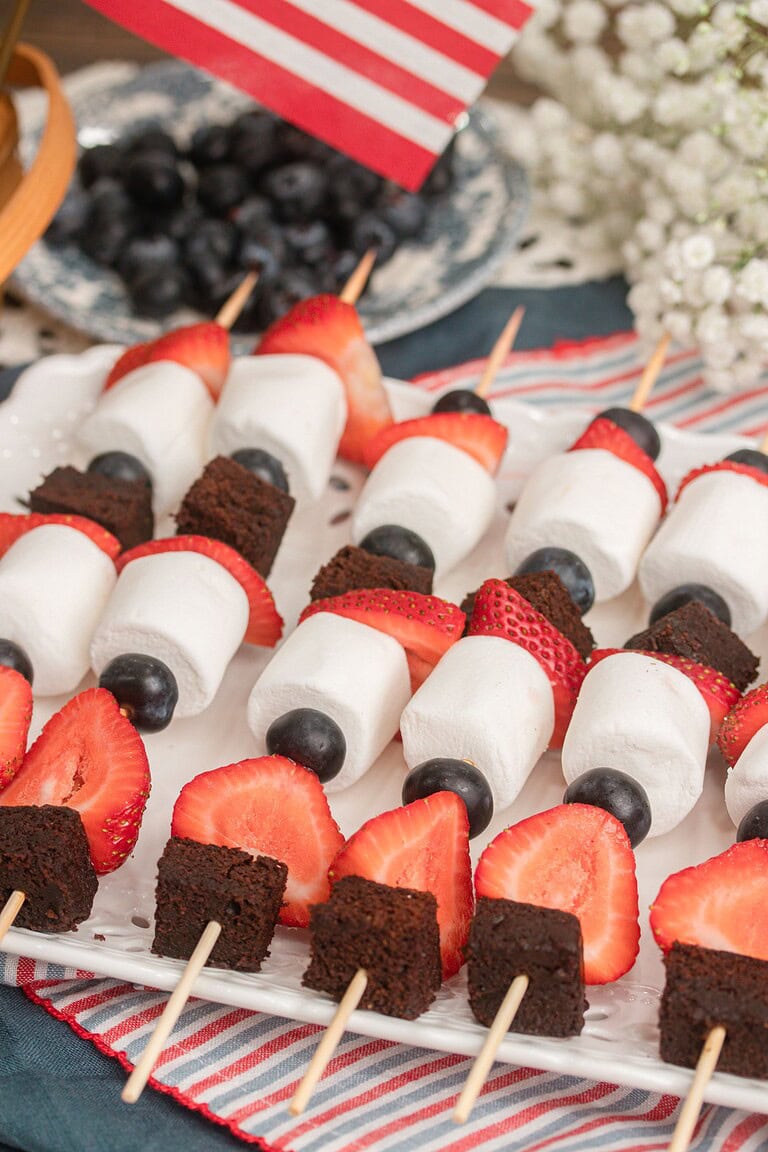 Close-up of red white and blue fruit kabobs with brownie bites, marshmallows, strawberries, and blueberries on a striped napkin
