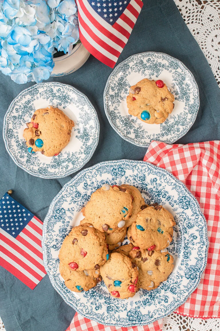Overhead view of red white and blue sourdough chocolate chip cookies stacked on floral plates with an American flag and hydrangeas