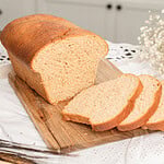 A whole loaf of Fresh Milled Flour Sandwich Bread on a wooden cutting board with several slices cut showing a soft even crumb, dried wheat stalks in the foreground and a farmhouse kitchen with white cabinets in the background