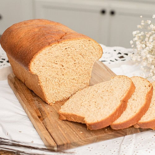 A whole loaf of Fresh Milled Flour Sandwich Bread on a wooden cutting board with several slices cut showing a soft even crumb, dried wheat stalks in the foreground and a farmhouse kitchen with white cabinets in the background