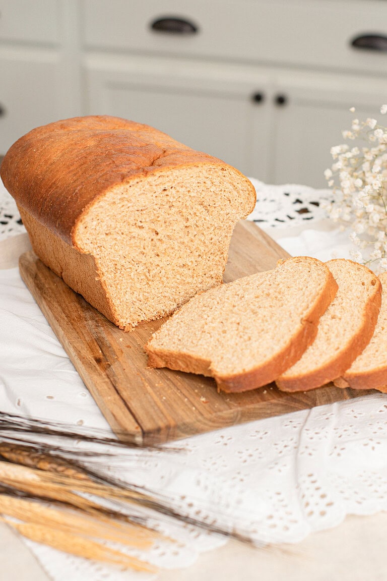 A whole loaf of Fresh Milled Flour Sandwich Bread on a wooden cutting board with several slices cut showing a soft even crumb, dried wheat stalks in the foreground and a farmhouse kitchen with white cabinets in the background