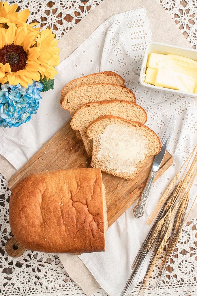 Four slices of Fresh Milled Flour Sandwich Bread fanned out on a wooden board, one slice spread with butter, next to a butter dish, a vintage knife, sunflowers, blue hydrangeas, and dried wheat stalks