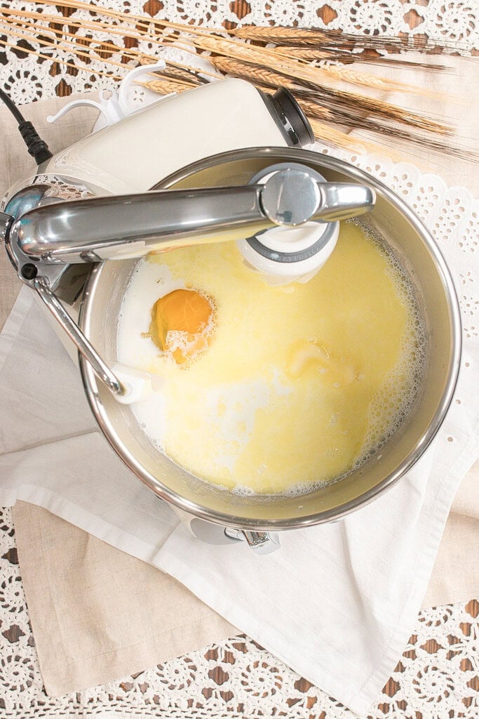 Overhead view of a stand mixer bowl with scalded milk, a cracked egg, and melted butter combined for the wet ingredients of sandwich bread dough