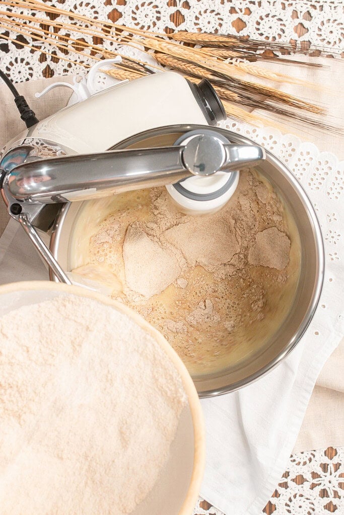 Freshly milled flour being added to the wet ingredients in a stand mixer bowl, with a bowl of flour resting nearby ready to be poured in