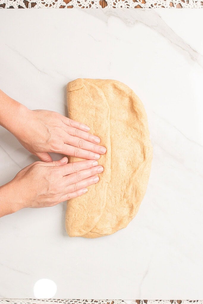 Hands folding fresh milled flour dough into thirds on a marble countertop as the first step in shaping the loaf