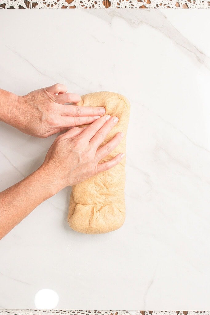 Hands pinching and sealing the seam of shaped sandwich bread dough on a marble countertop before placing it in the loaf pan