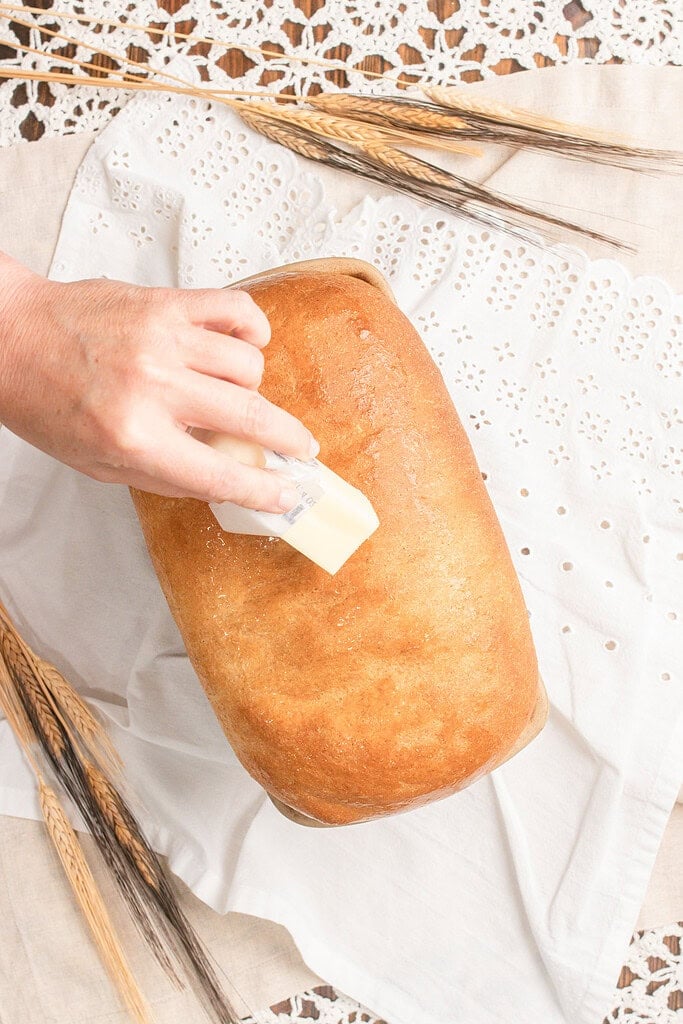 A hand brushing melted butter over the crust of a freshly baked golden Fresh Milled Flour Sandwich Bread loaf, still in the pan