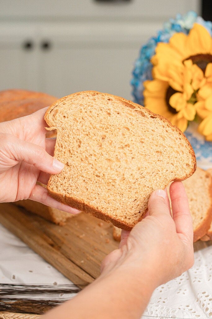 Close up of hands holding a soft slice of Fresh Milled Flour Sandwich Bread showing an even tender crumb, with a sliced loaf, sunflowers, and blue hydrangeas blurred in the background