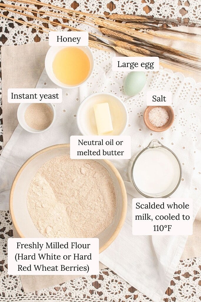 Overhead flat lay of sandwich bread ingredients including honey, a large egg, instant yeast, neutral oil or melted butter, salt, scalded whole milk cooled to 110 degrees, and a bowl of freshly milled hard white or hard red wheat flour, arranged on linen with dried wheat stalks