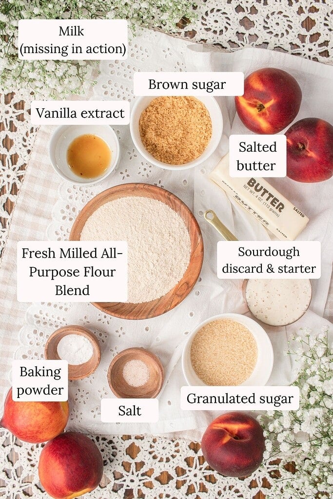 Overhead ingredient photo for sourdough peach cobbler with fresh milled flour featuring fresh peaches, fresh milled all-purpose flour blend, sourdough starter and discard, brown sugar, granulated sugar, butter, vanilla extract, baking powder, salt, and milk on a vintage linen backdrop.
