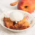 Sourdough peach cobbler with fresh milled flour topped with vanilla ice cream in a floral bowl, fresh peaches and cinnamon stick in background