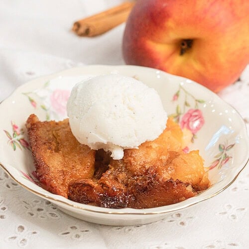 Sourdough peach cobbler with fresh milled flour topped with vanilla ice cream in a floral bowl, fresh peaches and cinnamon stick in background