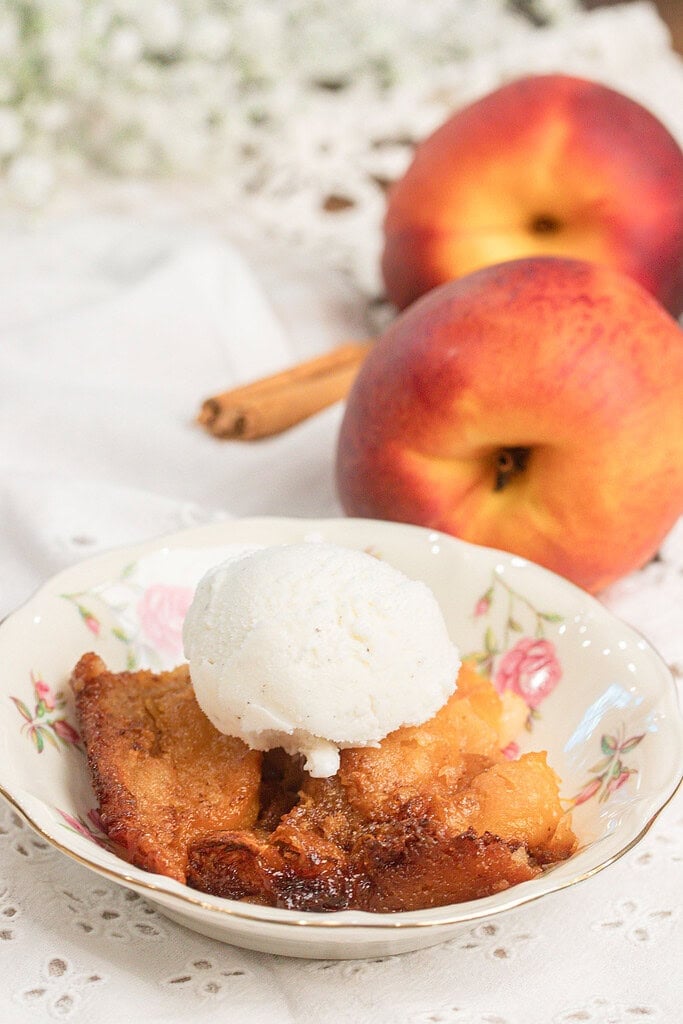 Sourdough peach cobbler with fresh milled flour topped with vanilla ice cream in a floral bowl, fresh peaches and cinnamon stick in background