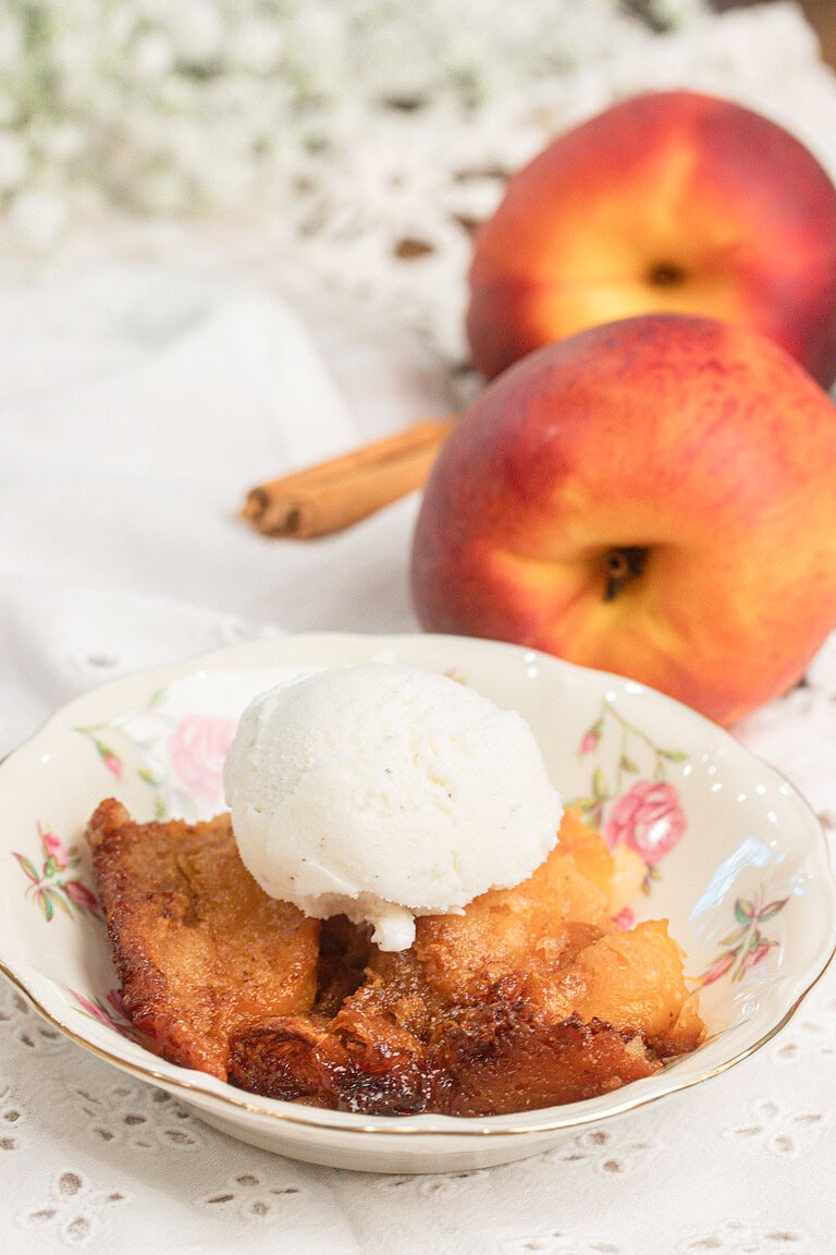 Sourdough peach cobbler with fresh milled flour topped with vanilla ice cream in a floral bowl, fresh peaches and cinnamon stick in background
