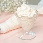 A glass dessert cup filled with swirled stabilized whipped cream, garnished with a tall piped peak, resting beside a piping bag on a soft pink linen with baby's breath in the background