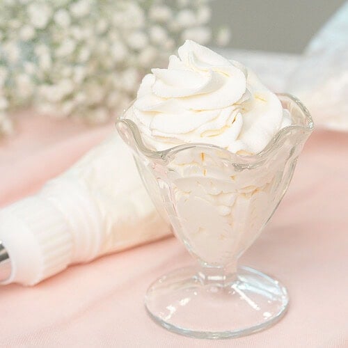 A glass dessert cup filled with swirled stabilized whipped cream, garnished with a tall piped peak, resting beside a piping bag on a soft pink linen with baby's breath in the background