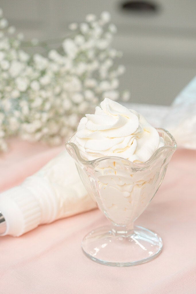 A glass dessert cup filled with swirled stabilized whipped cream, garnished with a tall piped peak, resting beside a piping bag on a soft pink linen with baby's breath in the background