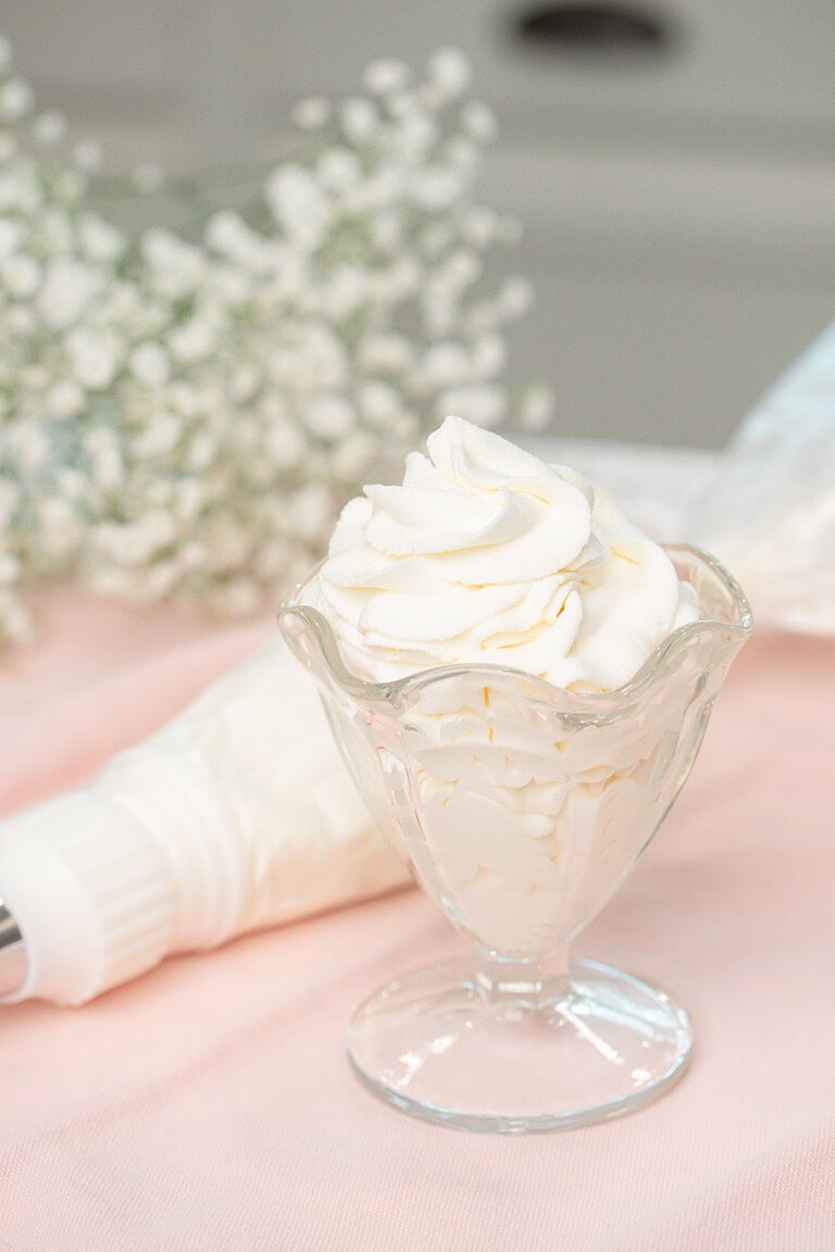 A glass dessert cup filled with swirled stabilized whipped cream, garnished with a tall piped peak, resting beside a piping bag on a soft pink linen with baby's breath in the background