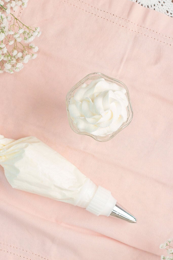 Overhead close up of a glass dessert dish filled with tightly piped stabilized whipped cream rosettes next to a filled piping bag on pink fabric