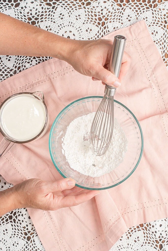 Hands whisking powdered sugar in a glass bowl with a measuring cup of heavy whipping cream sitting nearby, preparing the dry mixture for stabilized whipped cream