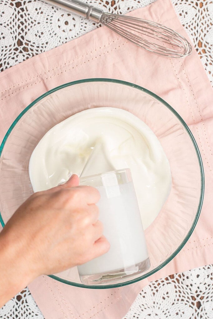 A hand pouring cornstarch into a glass bowl of powdered sugar while whisking the dry ingredients together for stabilized whipped cream