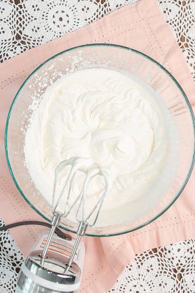 Freshly whipped cream in a glass bowl showing soft swirled peaks, with a hand mixer resting in the bowl after whipping to soft peaks