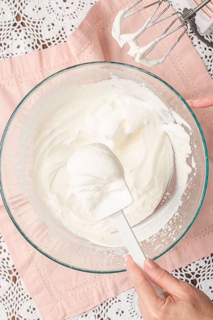 Stabilized whipped cream lifted on a rubber spatula showing thick glossy stiff peaks, with more whipped cream clinging to the beaters of a hand mixer above the bowl