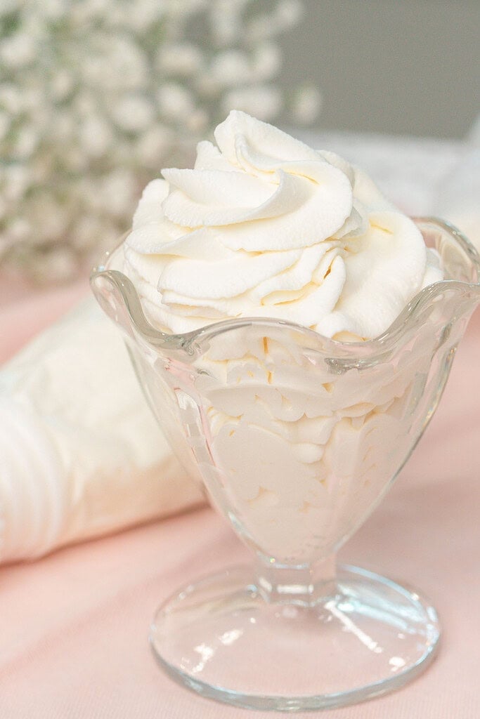 Close up of a glass dessert dish overflowing with piped stabilized whipped cream in soft rosette swirls, with a piping bag blurred in the background