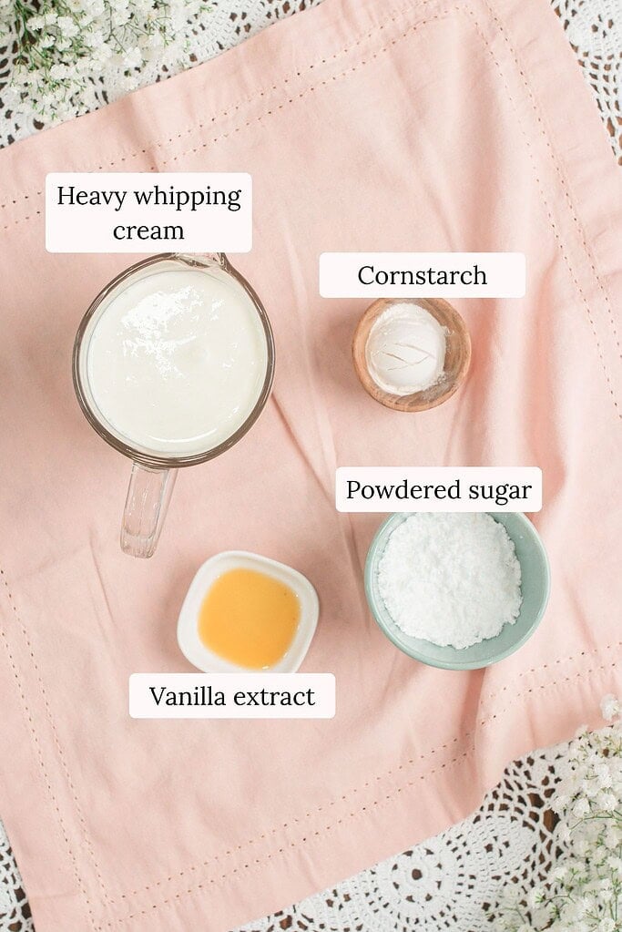 Overhead flat lay of stabilized whipped cream ingredients including a measuring cup of heavy whipping cream, a small dish of cornstarch, a bowl of powdered sugar, and a dish of vanilla extract on a pink linen