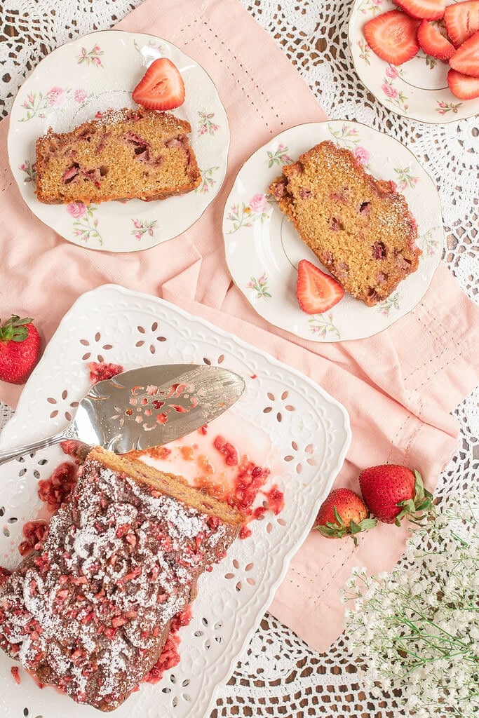 Two slices of Fresh Milled Flour Strawberry Bread plated on vintage floral china with a fresh strawberry half on top, powdered sugar dusted over the sliced loaf on a scalloped white platter in the foreground, surrounded by loose strawberries and baby's breath on pink linen