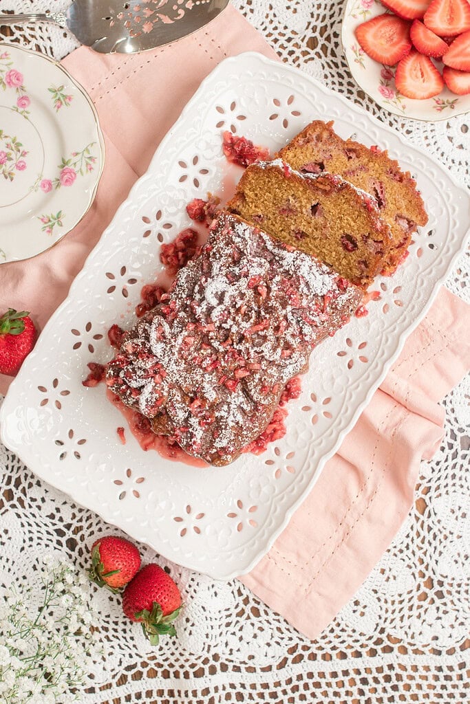 Sliced Fresh Milled Flour Strawberry Bread on a white scalloped platter, dusted with powdered sugar and drizzled with strawberry glaze, with fresh strawberries and a cake server nearby