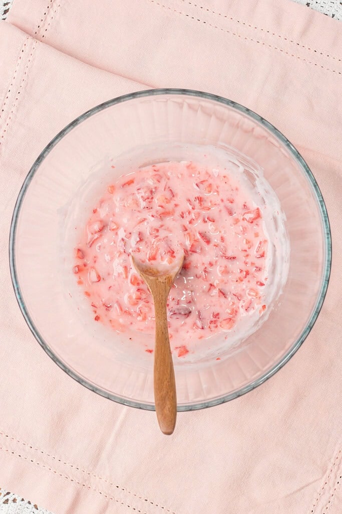 Strawberry buttermilk glaze being stirred in a glass bowl with a wooden spoon, showing diced strawberries folded into a pink glaze