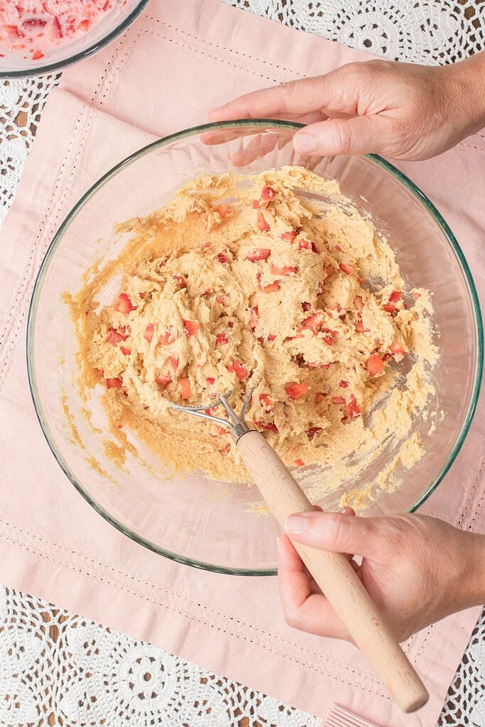 Hands folding fresh diced strawberries into fresh milled flour quick bread batter in a glass mixing bowl using a wooden spoon.