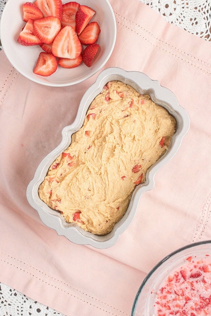 Strawberry bread batter filled into a decorative floral loaf pan, with a bowl of sliced strawberries and the strawberry glaze mixture nearby