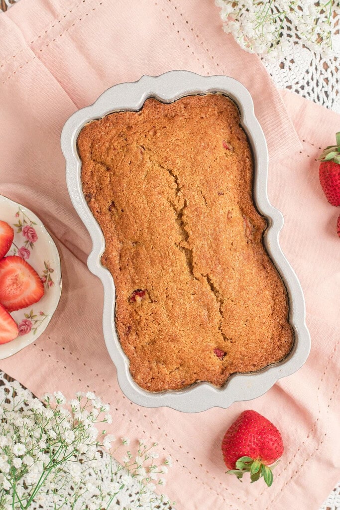 Baked Fresh Milled Flour Strawberry Bread in a floral shaped loaf pan, golden brown with a cracked top, resting on pink linen surrounded by strawberries and baby's breath