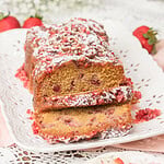Close up of sliced Fresh Milled Flour Strawberry Bread on a white platter, showing a moist crumb studded with strawberries and a powdered sugar dusted glaze, with baby's breath and fresh strawberries in the background