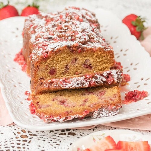 Close up of sliced Fresh Milled Flour Strawberry Bread on a white platter, showing a moist crumb studded with strawberries and a powdered sugar dusted glaze, with baby's breath and fresh strawberries in the background