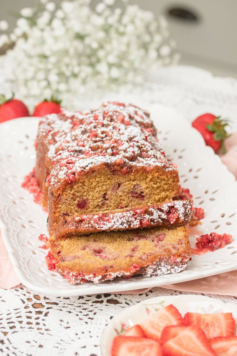 Close up of sliced Fresh Milled Flour Strawberry Bread on a white platter, showing a moist crumb studded with strawberries and a powdered sugar dusted glaze, with baby's breath and fresh strawberries in the background